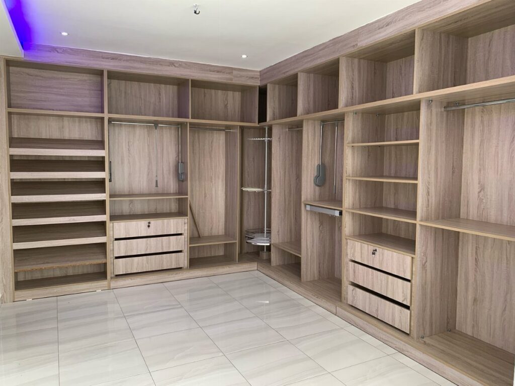 L-shaped walk-in closet with light wood shelving and built-in drawers on white tile floors