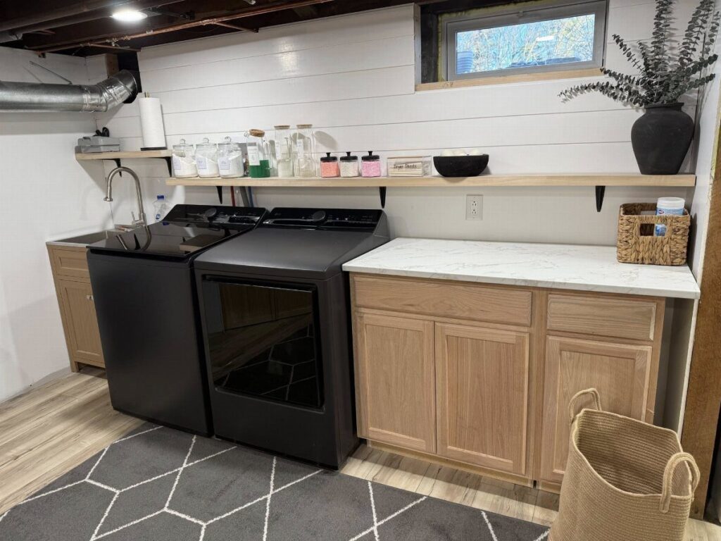 Black washer and dryer with light wood cabinets and white shiplap walls.