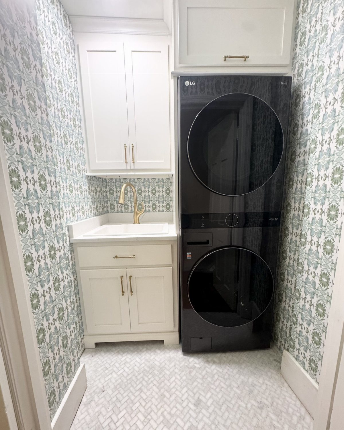 Small laundry room with blue floral wallpaper, white cabinets, and black stacked laundry machines.