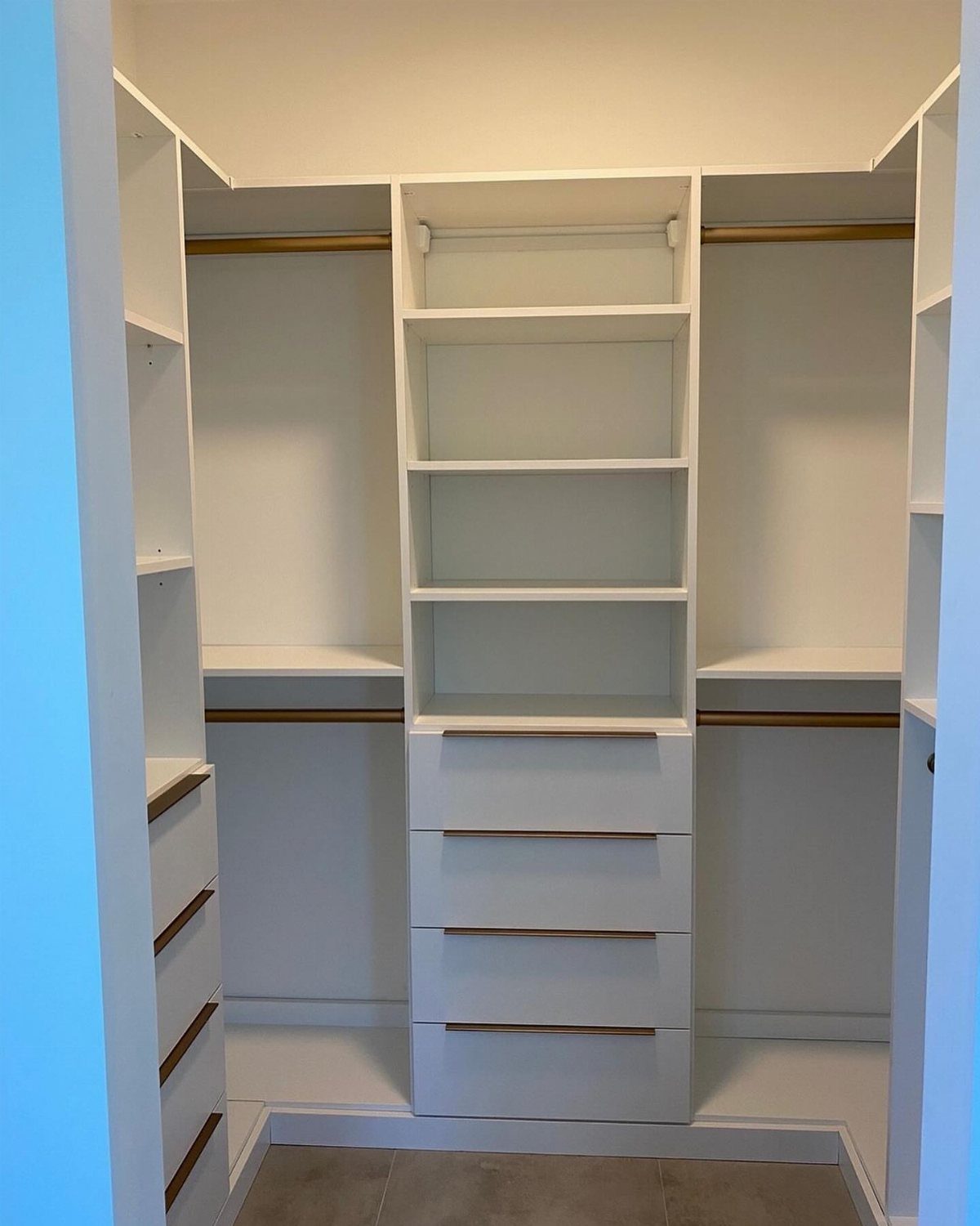 White walk-in closet with gold hanging rods, central shelving, and four drawers.