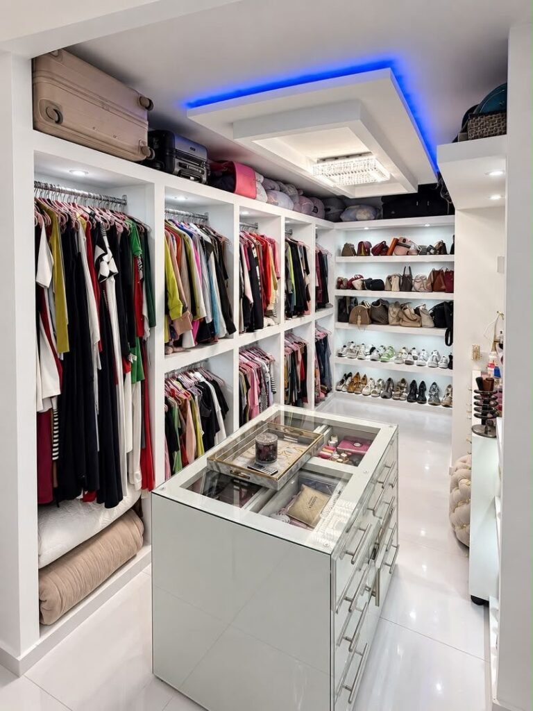 White walk-in closet with organized clothing, a central glass-top island, and blue ceiling lighting.