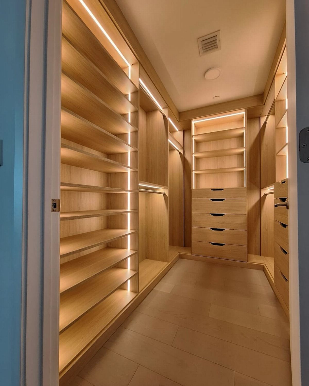 Walk-in closet with light wood shelving illuminated by integrated warm LED light strips.